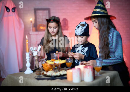 Cute little friends in fancy costumes gathered together at Halloween home party and stuffing their faces with tasty sweet snacks, interior of decorate Stock Photo