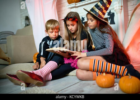 Group of cute kids wearing Halloween costumes gathered together at living room decorated for holiday and reading short scary stories Stock Photo