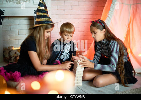 Little guests of Halloween party: group of cute children enjoying delicious fried marshmallows while sitting on floor and playing board game Stock Photo