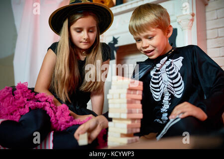 Handsome little boy and his pretty elder sister wearing Halloween costumes sitting on floor of living room and playing board game with enthusiasm Stock Photo