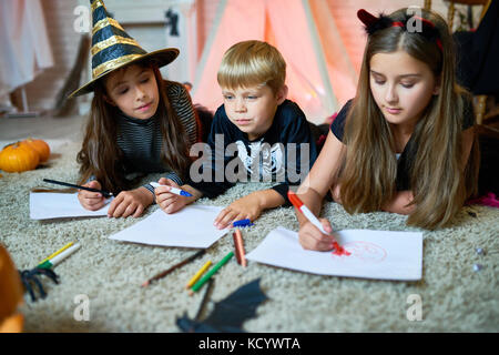 Cute little friends wearing fancy costumes lying on cozy carpet at living room decorated for celebrating Halloween and drawing with colorful felt-tip  Stock Photo