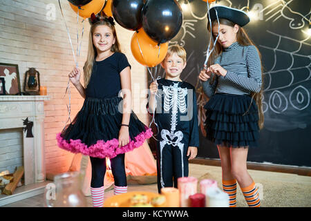 Group portrait of smiling little friends wearing fancy costumes holding bunches of balloons in hands while posing for photography, interior of living  Stock Photo