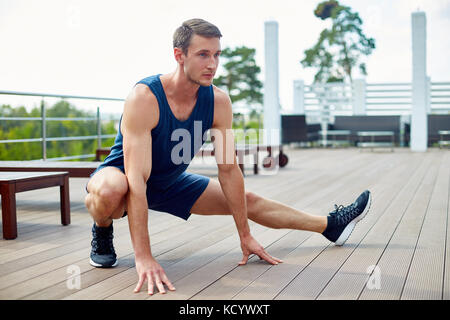 Handsome young sportsman doing side lunge while having training on spacious wooden terrace, picturesque view on background Stock Photo