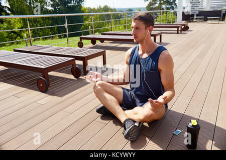 Sporty young man sitting in lotus position and listening to music while meditating on spacious terrace with breathtaking view Stock Photo