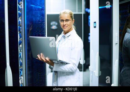 Portrait of young woman wearing lab coat and holding laptop smiling at camera while working with supercomputer in server room Stock Photo