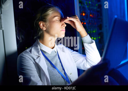 Portrait of beautiful young woman sitting on floor in dark server room using laptop while working with supercomputer at night, rubbing forehead fighti Stock Photo