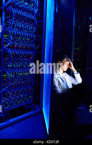 Portrait of exhausted young woman sitting on floor in dark server room using laptop while working with supercomputer at night Stock Photo