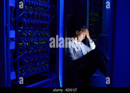 Portrait of tired young woman sitting on floor in dark server room using laptop while working with supercomputer at night Stock Photo