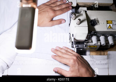 Above view closeup of tailors hands leading white fabric through serger machine working in lamplight at night Stock Photo