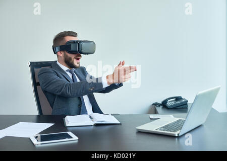 Portrait of handsome modern businessman using VR glasses playing shooter game sitting at desk in office Stock Photo
