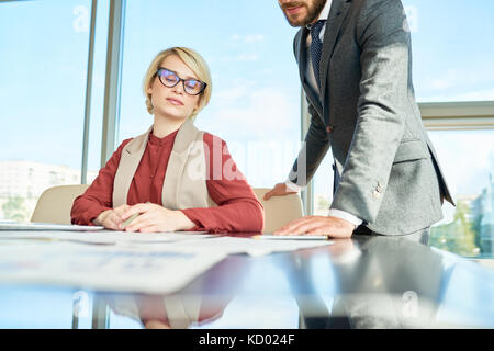 Portrait of modern successful businesswoman sitting at conference table in office against window and talking to assistant discussing documentation Stock Photo