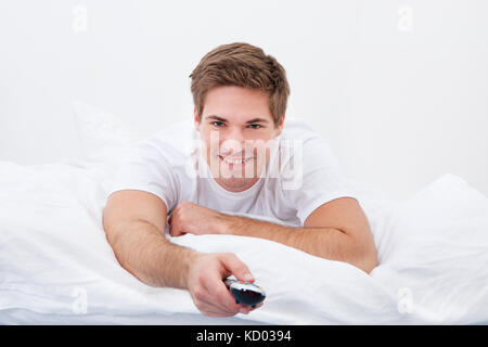 Young Man Lying On Bed Changing Channel With Remote Control Stock Photo