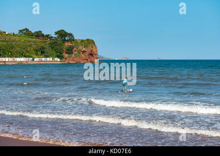 Paddle-boarder heading out to sea at Goodrington Sands, Paignton, Devon. UK. Beach huts and red cliffs at Roundham Head with boat in the distance Stock Photo