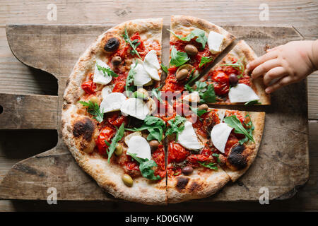 Sliced pizza on chopping board, child's hand reaching for pizza slice, overhead view Stock Photo