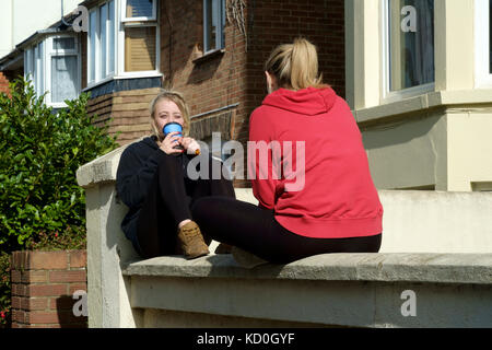 two young female students sat relaxing on a wall outside of their accommodation chatting and drinking coffee england uk Stock Photo