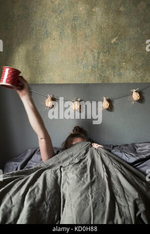 Woman in bed, hiding under covers, holding mug in air Stock Photo