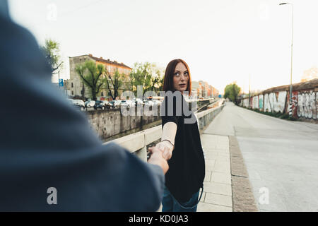 Over shoulder view of young woman holding boyfriend's hand by city canal Stock Photo