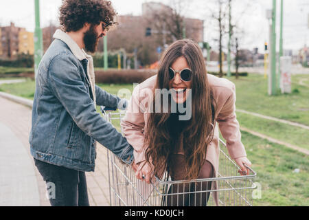 Young couple outdoors, man pushing woman along in shopping trolley Stock Photo