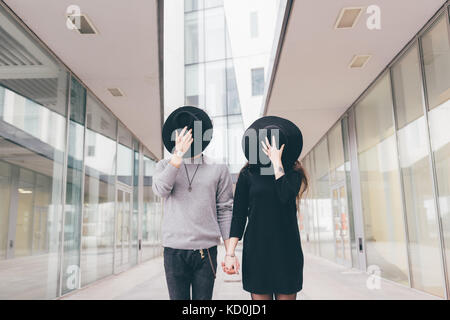 Portrait of young couple in urban environment, holding hands, covering faces with hats Stock Photo
