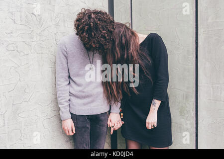 Young couple outdoors, standing against wall, holding hands, hair covering their faces, Stock Photo