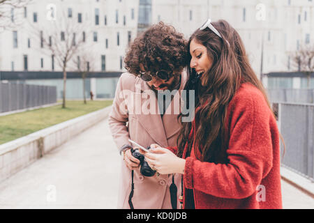 Young couple standing outdoors, looking at smartphone, laughing Stock Photo