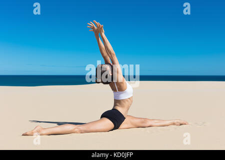 Side view of woman on beach doing the splits, arms raised in yoga position Stock Photo