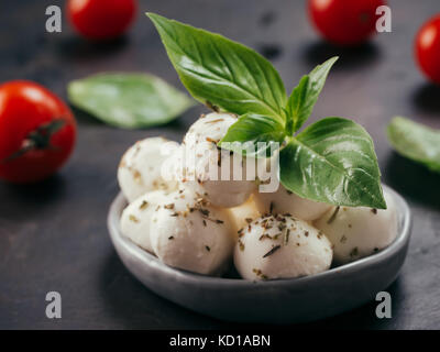 Close up view of small mozzarella cheese marinated with herbs. Mozarella balls with fresh basil and cherry tomatoes on black concrete background. Low  Stock Photo
