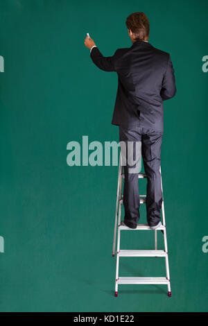 Rear View Of A Businessman Standing On Ladder Writing On Chalkboard Stock Photo