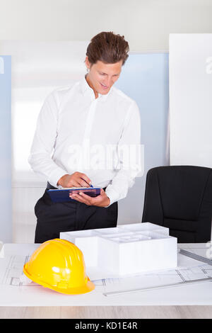 Male Architect Looking At Architectural Model Writing On Clipboard Stock Photo