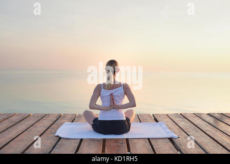Rear view of young woman sat on towel practicing yoga by sea at sunset Stock Photo