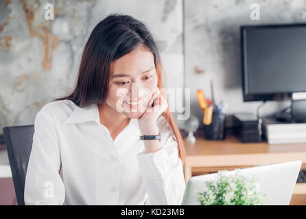 Asian businesswoman looking at laptop computer and smiling face and rest chin on hand with happy emotion from good news of business success in home of Stock Photo