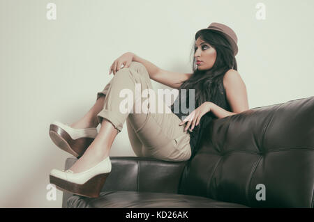 Side vierw portrait of a young Indian woman wearing a hat sitting on a leather sofa looking away Stock Photo