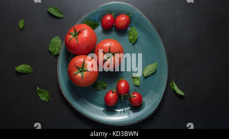 Ripe tomatoes and mint leaves in a plate. Big and small sized tomatoes in a ceramic plate on black  background. Stock Photo