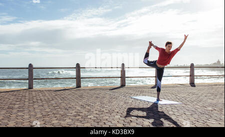 Woman stretching during yoga on road along the sea. Fit woman workout outdoors, doing Natarajasana, Dancer king pose on seaside promenade. Stock Photo