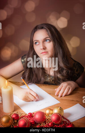 Beautiful girl with blue eyes, thinking hopeful of what to write to Santa, at candlelight, surrounded by colorful globes and Christmas lights. Stock Photo