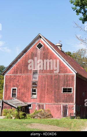 Rustic old red barn in disrepair with grass in the foreground and blue sky above.  A weathervane sits on the roof of the barn. Stock Photo