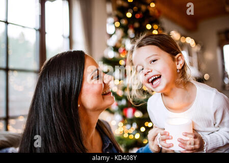 Beautiful young mother with little daughter at home by the Christmas tree together. Girl holding burning candle. Stock Photo