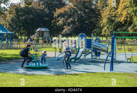 Young parents and their two small children in a playground / play area, on a warm sunny day, early autumn. Gunnersbury Park, West London, England, UK. Stock Photo