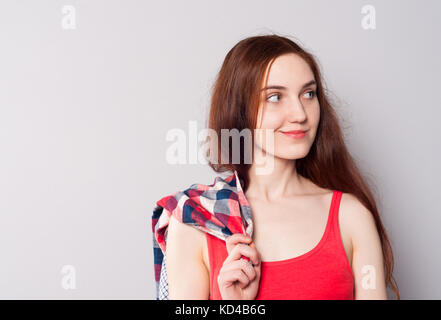 Young pretty red-haired girl in a T-shirt holding a trendy checkered shirt, waiting for someone, looking away and smiling on a light gray background Stock Photo