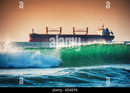 Sun setting at the sea with sailing cargo ship and crashing waves near the harbour Stock Photo