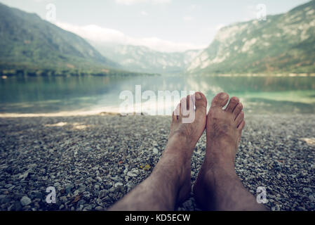 Summer holiday by the lake, barefoot adult caucasian male relaxing on beach pebbles and looking into water and mountains in the distance. Enjoying vac Stock Photo