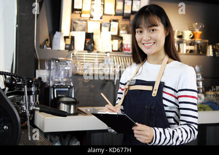 Portrait of smiling asian barista listing food. Young asian woman barista in her shop. Cafe restaurant service, food and drink industry concept. Stock Photo