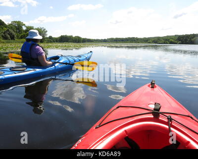 Two friends kayaking on a lake in red and blue canoes.  The bow of the red kayak is visible as is the middle-aged woman in a blue kayak. Stock Photo