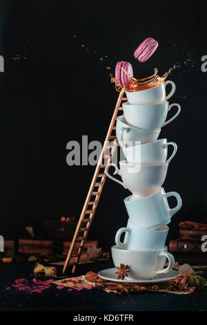 Tower of white coffee cups with a ladder, macaroons, splash and coffee drops on a dark background. Indulgence concept. Action food photography. Stock Photo