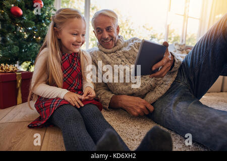 Grandfather and granddaughter sitting on floor and looking at digital tablet during christmas. Man with his little girl having a video on digital tabl Stock Photo