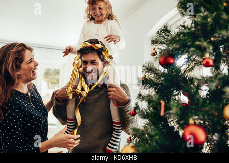 Happy young couple with their daughter celebrating Christmas. Little girl sitting on her father's shoulders and having fun. Stock Photo