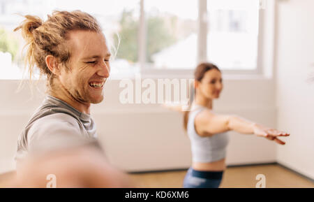 Smiling young man doing warrior pose in yoga class with female student. Happy fitness male practicing yoga at gym class. Stock Photo