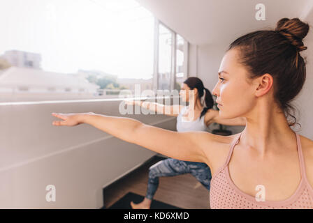 Women doing yoga in warrior pose at studio. Young fitness females working out indoors. Stretching yoga workout in gym class. Stock Photo