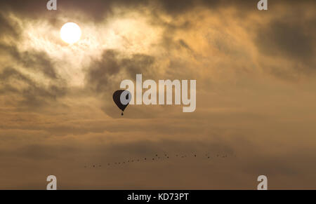Isolated, UK hot air balloon silhouetted in autumn sunset sky, late sun shining brightly through fluffy clouds. Concept getaway, social distancing. Stock Photo