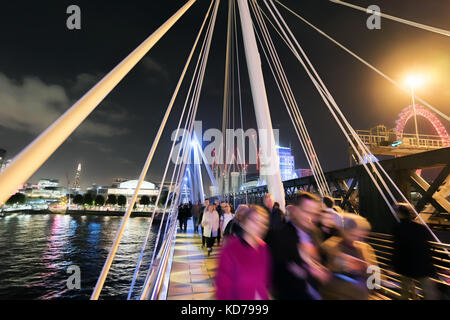 People walking along the Hungerford footbridge against the backdrop of the South Bank at night Stock Photo